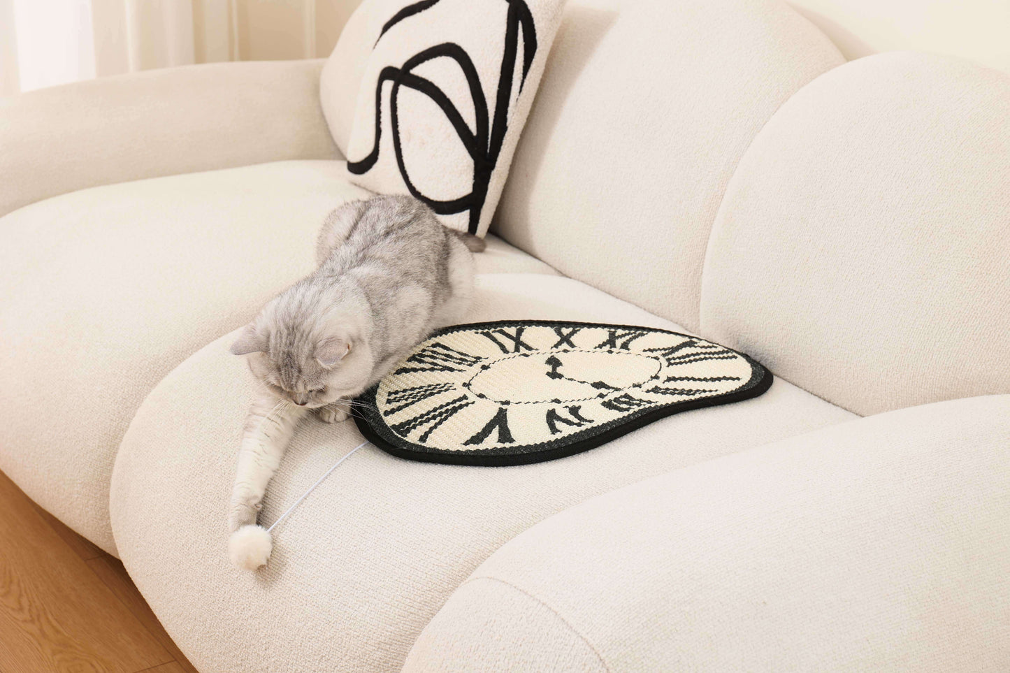 Cat playing with a pompom on a Melting Clock Cat Scratching Mat on a beige couch.
